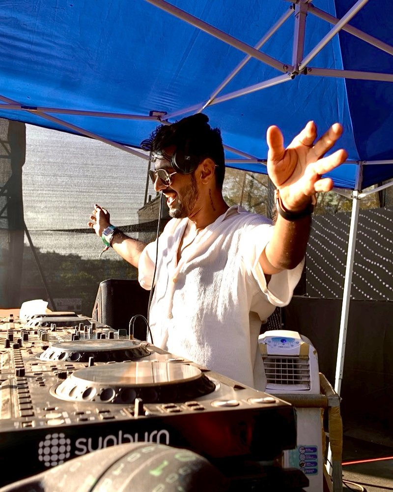 Aryan with arms open behind the decks at the Sunburn booth in afternoon sun